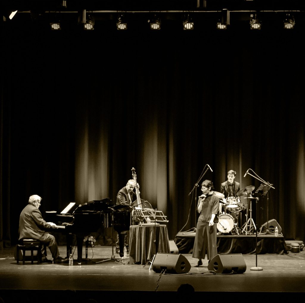 A Quartet - a four-piece band, consisting of a pianist playing a baby grand piano, with a double bass player, a drummer and a vocalist performing on stage.