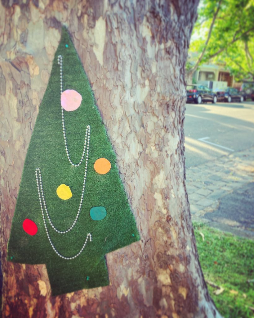 A cutout green artificial grass shaped like a Christmas tree adhered to a tree trunk. The Christmas tree cutout is adorned with circular cut-out coloured papers and strings. 